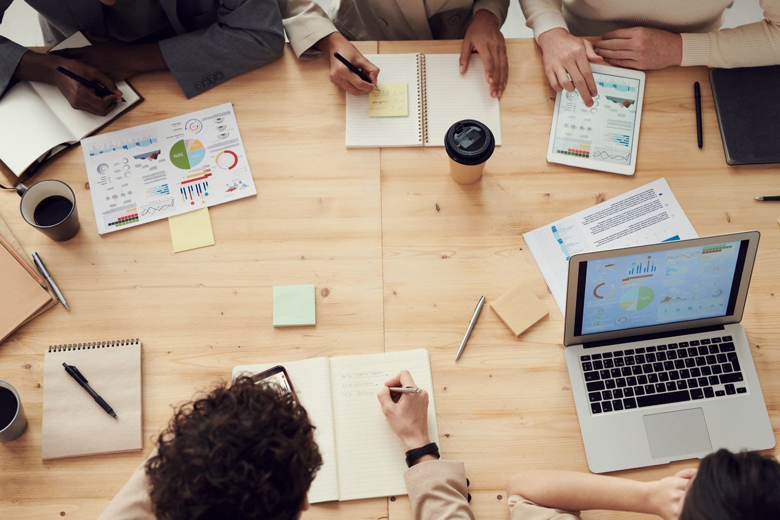 A group of people meeting around a table with printed reports showing charts, a tablet and a laptop displaying figures